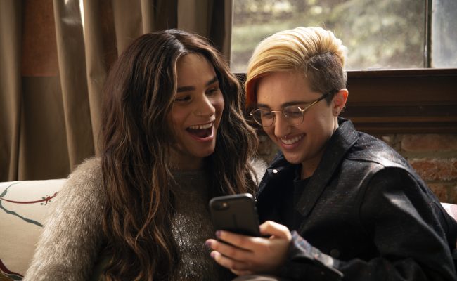 A transfeminine non-binary person and transmasculine gender-nonconforming person looking at a phone and laughing