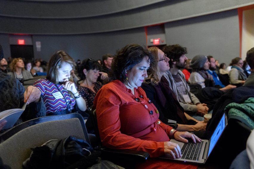 Woman sitting in an auditorium among many other people, looking down at her laptop and working.